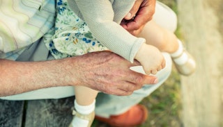 A young child sitting in the lap of a grandparent and holding its hand