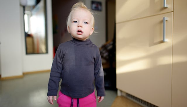 A blonde female toddler wearing a black shirt with a curious facial expression standing in a room