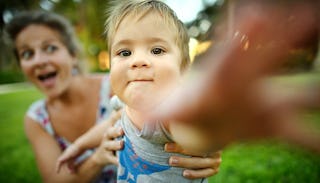 A blurred smiling mother in the background holding her baby while reaching her hand out in the direc...