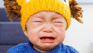 A crying baby wearing a blue shirt with white dots and a yellow wool hat