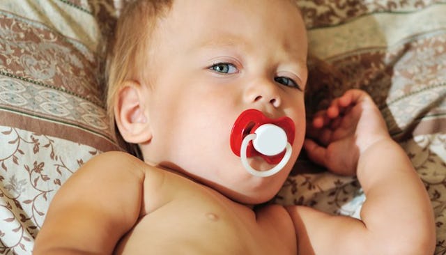 A baby lying on a bed with a red-white pacifier in his mouth