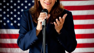 Woman wearing a suit speaking on the microphone with the flag of the United States in the background...