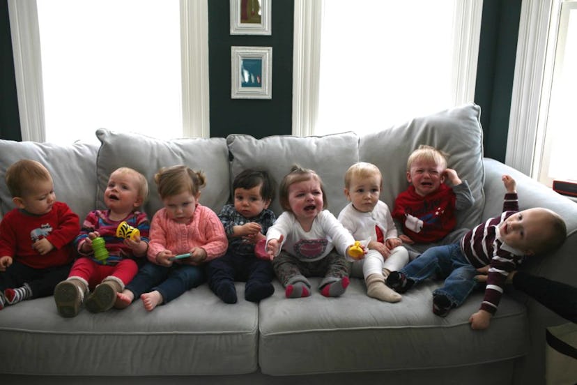 Eight toddlers sitting on a gray couch next to each other; some of them are crying, and some of them...