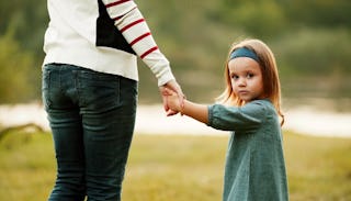 A mother is holding a young girl's hand while the girl is looking at the camera in a green dress and...