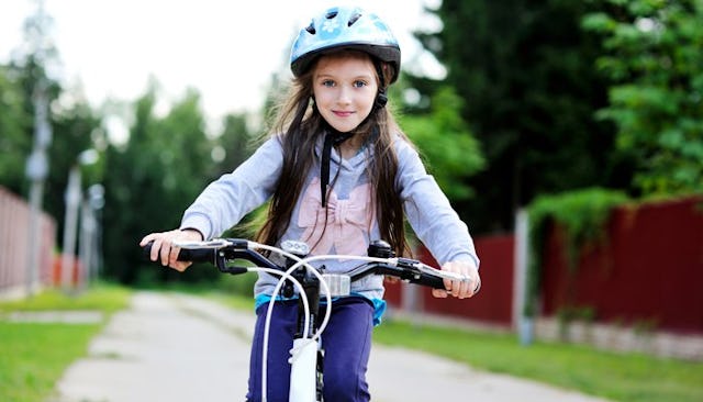 A brown-haired girl with blue eyes, in a blue hoodie with a pink bow, riding a bike down the street