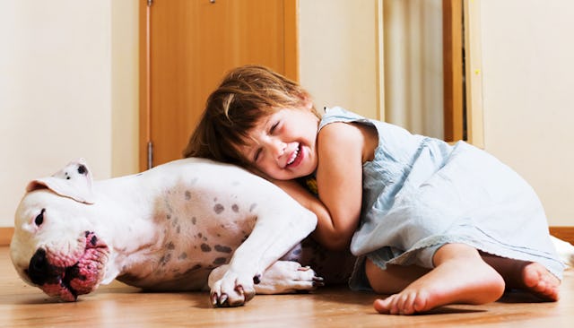 A smiling toddler in a grey linen dress hugging and lying on a white dog on the floor