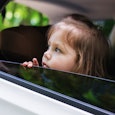 A girl with brown hair is looking through the window of a white car while being dropped off at schoo...