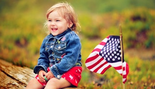 A toddler in red shorts and a blue denim jacket smiling while sitting on a bench next to a small Ame...
