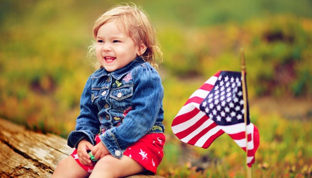 A toddler in red shorts and a blue denim jacket smiling while sitting on a bench next to a small Ame...