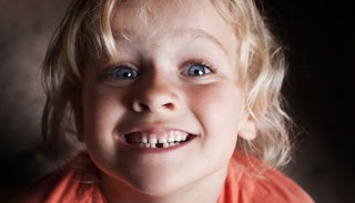 A blonde boy in an orange shirt, flashing a joyful smile, having lost one of his teeth, awaiting the...