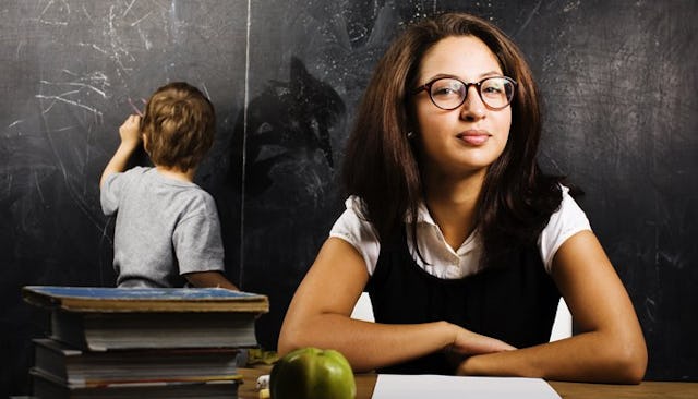 An elementary teacher sitting at the desk while a kid writes something on a blackboard behind her