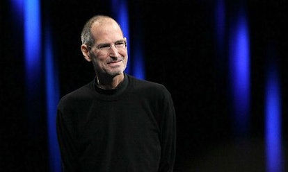 Steve Jobs wearing a black long sleeve shirt and glasses smiling with the background black and blue