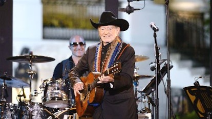 Willie Nelson wearing a cowboy hat and suit holding a guitar while playing with a band on stage