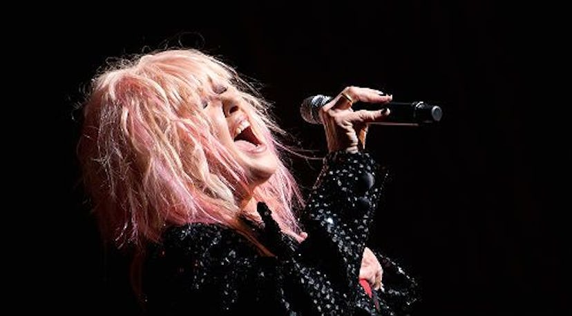 Cyndi Lauper with pink hair, wearing a black jacket and singing into a mic