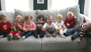 Eight toddlers sitting on a gray couch next to each other; some of them are crying, and some of them...