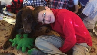 A kid with autism in a red long sleeve shirt wearing glasses leaned on a black dog with a group of p...