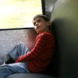 A boy in a red long-sleeved shirt with grey stripes and blue denim jeans sitting in a school bus loo...