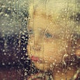 A little boy who has autism is looking through a window that has rain drops on it while looking sad 