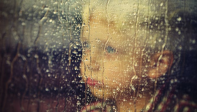 A little boy who has autism looking through a window that has rain drops on it while looking sad