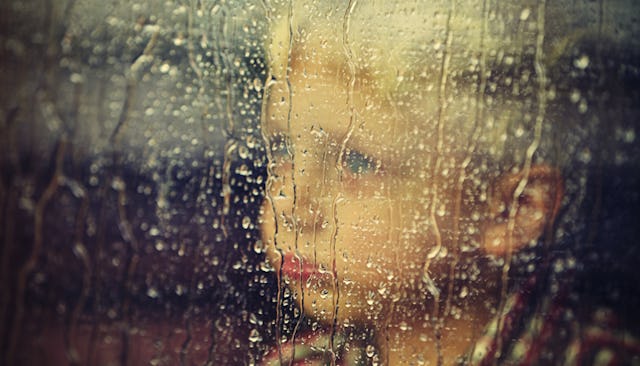 A little boy who has autism looking through a window that has rain drops on it while looking sad 