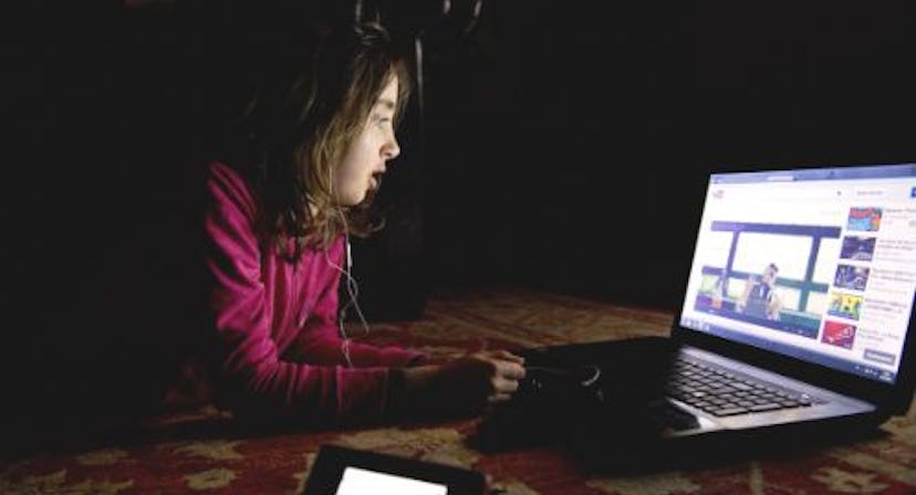 A girl in a burgundy hoodie in a dark room, lying on her stomach and looking into her laptop