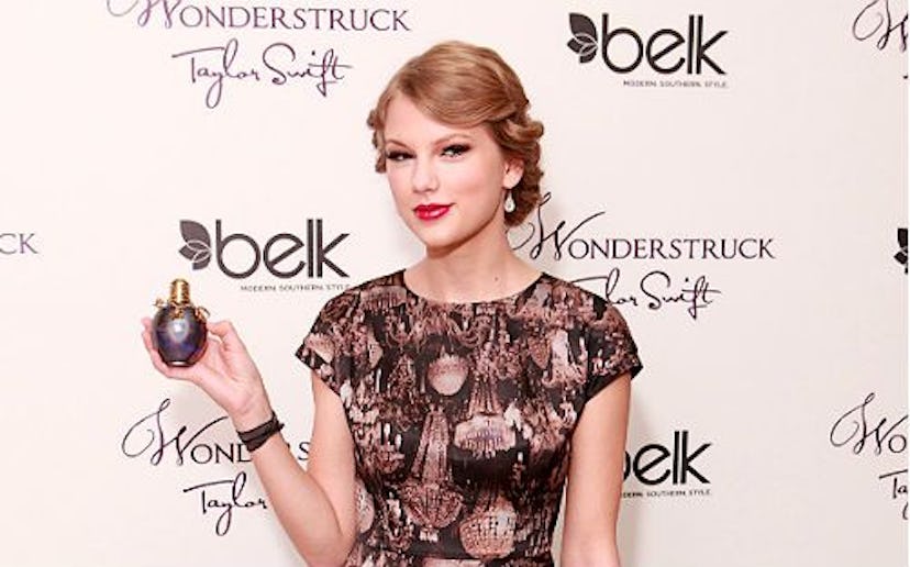 Taylor Swift, with her hair in an updo, wearing a shirt with a brown-golden print smiling and holdin...