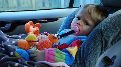 A baby in a car seat covered with a grey blanket with white dots holding orange plush toys