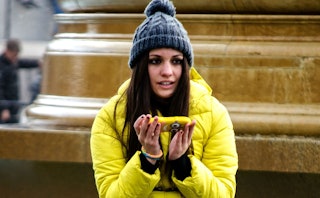 A brunette, long-haired girl with a gray knit in a yellow puffer jacket holding a Minion-themed phon...