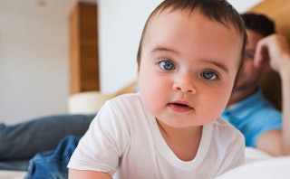 A blue-eyed baby in a white shirt lying on a bed with his father blurred in the background