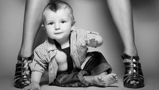 A baby sitting beside a pair of women's legs adorned with sleek black heels.