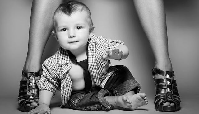 A baby sitting beside a pair of women's legs adorned with sleek black heels.
