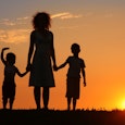 A single mother with curly hair in a midi dress between two children holding hands, watching a sunse...