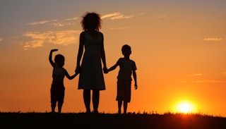 A single mother, wearing a midi dress, standing between two children holding hands, gazing at a suns...