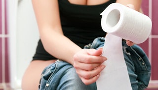 Woman sitting on the toilet holding a roll of toilet paper while dealing with hemorrhoids.