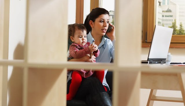 A working mom talking on a phone and looking at a laptop while she's holding a toddler on her lap