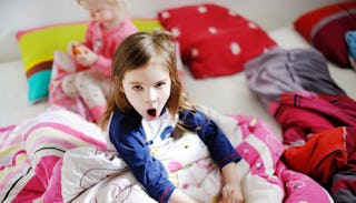 A toddler girl, looking upset, sitting on the bed in her pajamas, covered by her colorful blanket, w...