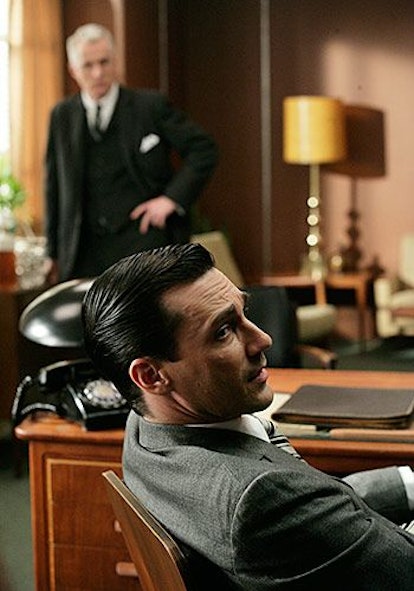 Don Draper sitting in his chair while Roger stands in the background in Mad Men 