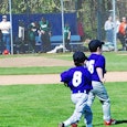 Three kids on a baseball field in blue shirts with the numbers 2, 8, and 16 on their backs, light bl...