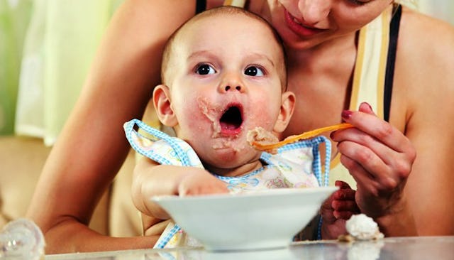 A happy mother feeding her baby while he is holding his hand inside the plate.