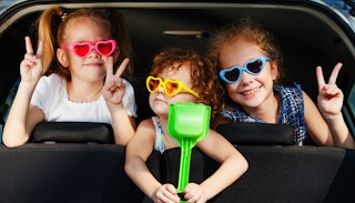 Three children wearing heart-shaped sunglasses in the backseat of a car on a road trip showing peace...