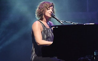 Sarah McLachlan wearing a grey dress singing into a microphone that is set on a piano