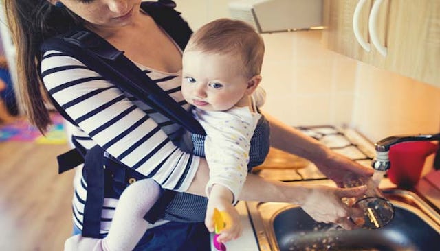 A stay-at-home mom caring for her baby in a baby carrier as she washes dishes.