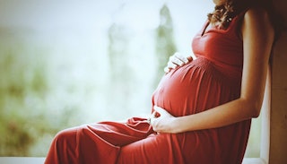 A young pregnant woman in a long red dress sitting alone next to a window with her hands on her bell...