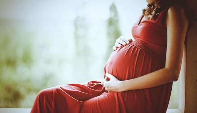 A young pregnant woman in a long red dress sitting alone next to a window with her hands on her bell...