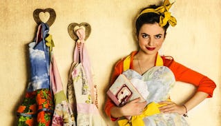 A woman dressed in 1960s style standing in the kitchen wearing an apron and a yellow ribbon on her h...