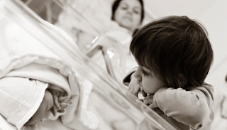 A toddler girl looking at her newborn sibling with their mother lying in a hospital bed in the backg...
