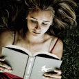 A blonde girl wearing a red tank top lying on the grass while reading a gray cover book that has a w...