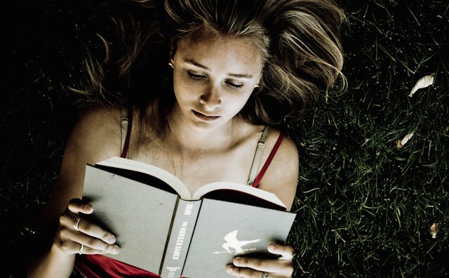 A blonde girl wearing a red tank top lying on the grass while reading a gray cover book that has a w...