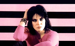 Joan Jett in a pink shirt with her hand on her head in front of a pink-and-black striped wall