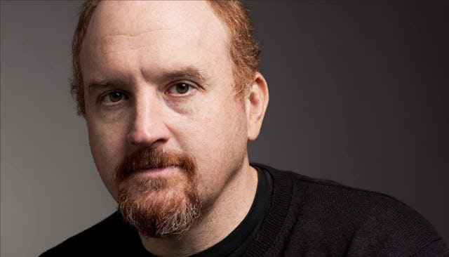 Louis C.K. wearing a black shirt while standing in front of a black background.